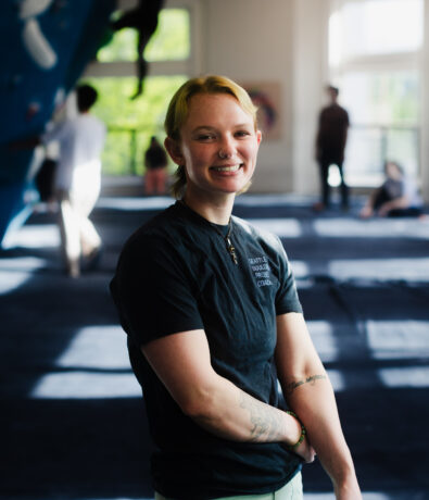 A person with short blond hair and tattoos smiles while standing in a brightly lit climbing gym in the University District, Seattle. Other people and colorful climbing walls are visible in the background.