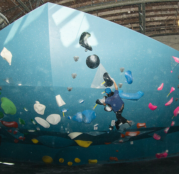 Indoor rock climbing gym in Salt Lake City features blue climbing walls filled with colorful holds under exposed industrial ceilings, with people watching and climbing. Enjoy waived initiation when you join the fun!.