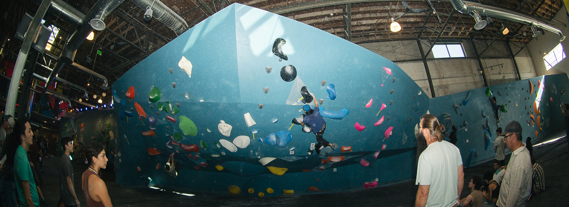 Indoor rock climbing gym in Salt Lake City features blue climbing walls filled with colorful holds under exposed industrial ceilings, with people watching and climbing. Enjoy waived initiation when you join the fun!.