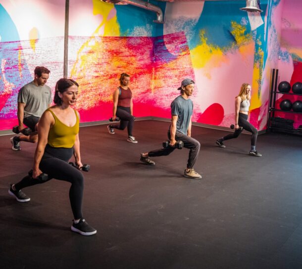 Five people in athletic wear perform walking lunges with dumbbells in a colorful, brightly painted gym studio with a black floor and wall-mounted shelves holding exercise balls in the background.