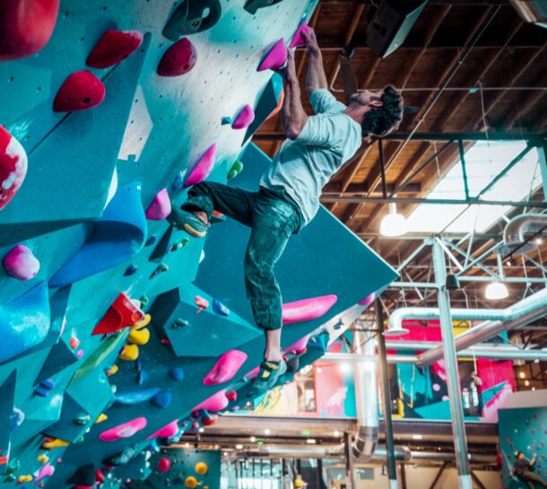 A person climbs an overhanging indoor bouldering wall, gripping colorful holds with both hands and feet. The gym has bright lighting, exposed ceiling beams, and vibrant climbing features.