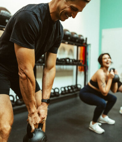 Two people exercising in a gym, holding kettlebells and smiling. The man in the foreground wears a black shirt and shorts, while the woman in the background wears a sports bra and leggings. Both appear to be enjoying the workout.