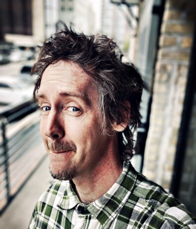 A man with tousled brown hair and a short beard, wearing a green and white plaid shirt, stands outdoors on a city sidewalk, looking at the camera with a slight smile. Urban buildings and parked cars are in the background.