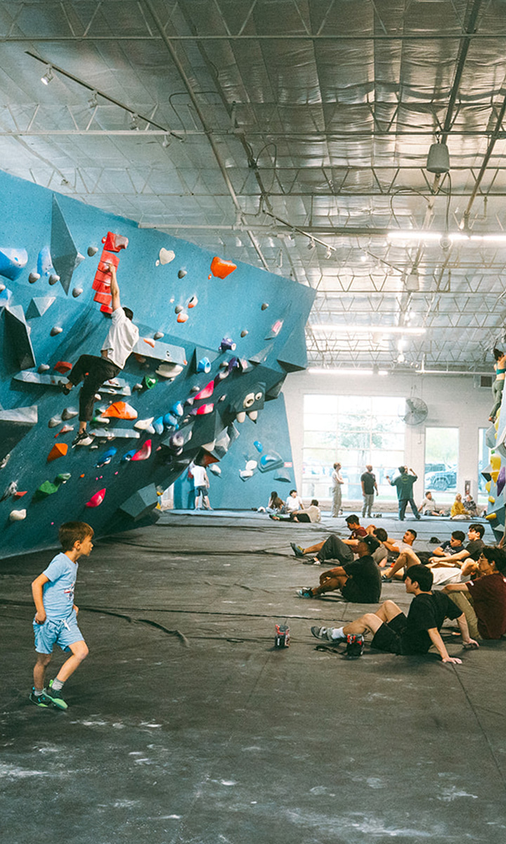 A busy indoor climbing gym with people bouldering on colorful climbing walls, others sitting or standing on padded floors, and a child walking in the foreground. Bright, spacious setting with natural light.