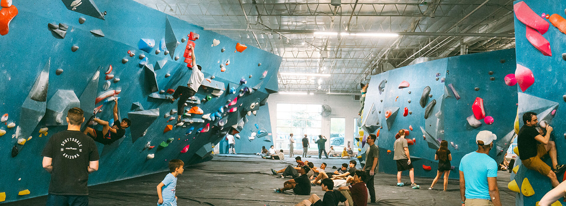 People of various ages are climbing and socializing in a large indoor bouldering gym with blue climbing walls, colorful holds, and soft mats covering the floor. Some are climbing while others sit or stand, watching and chatting.