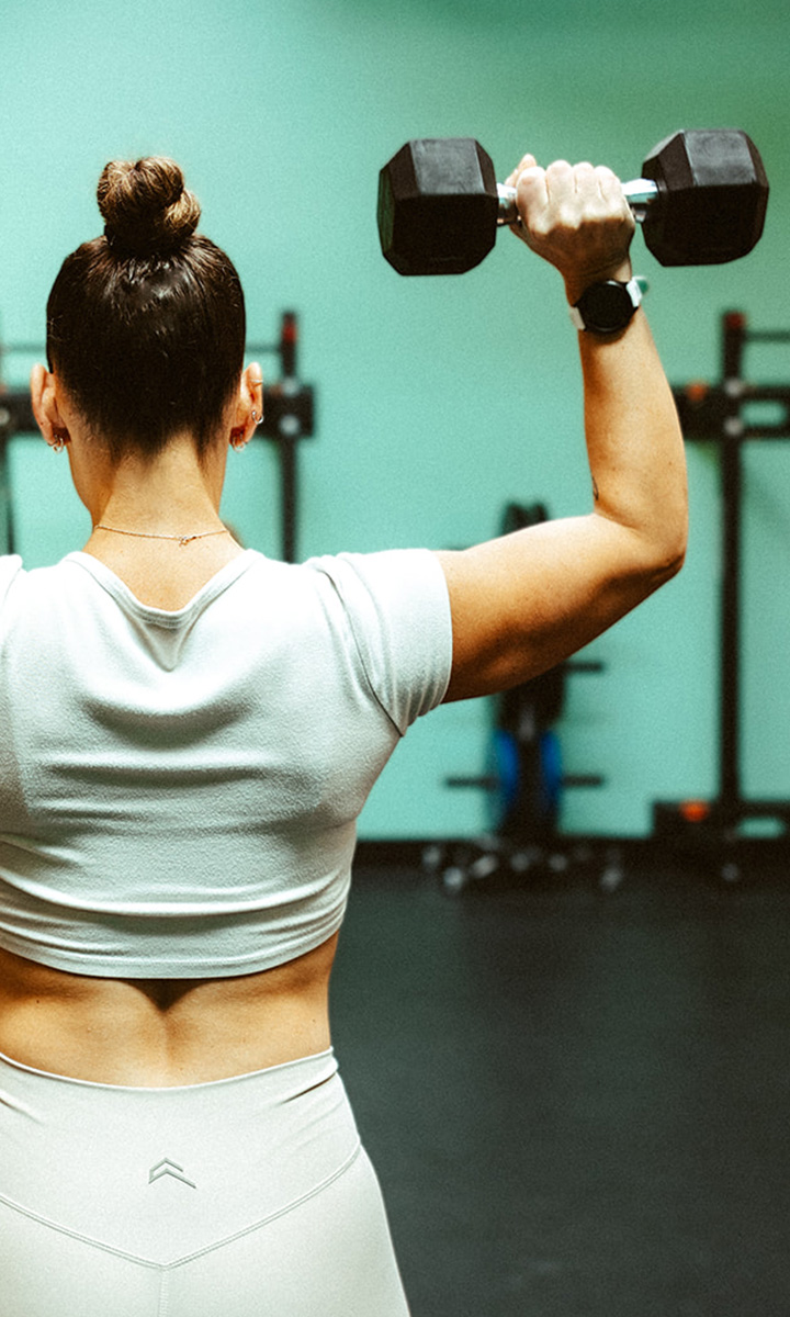 A woman with her hair in a bun, wearing a white crop top and leggings, lifts a dumbbell overhead in a gym, photographed from behind.
