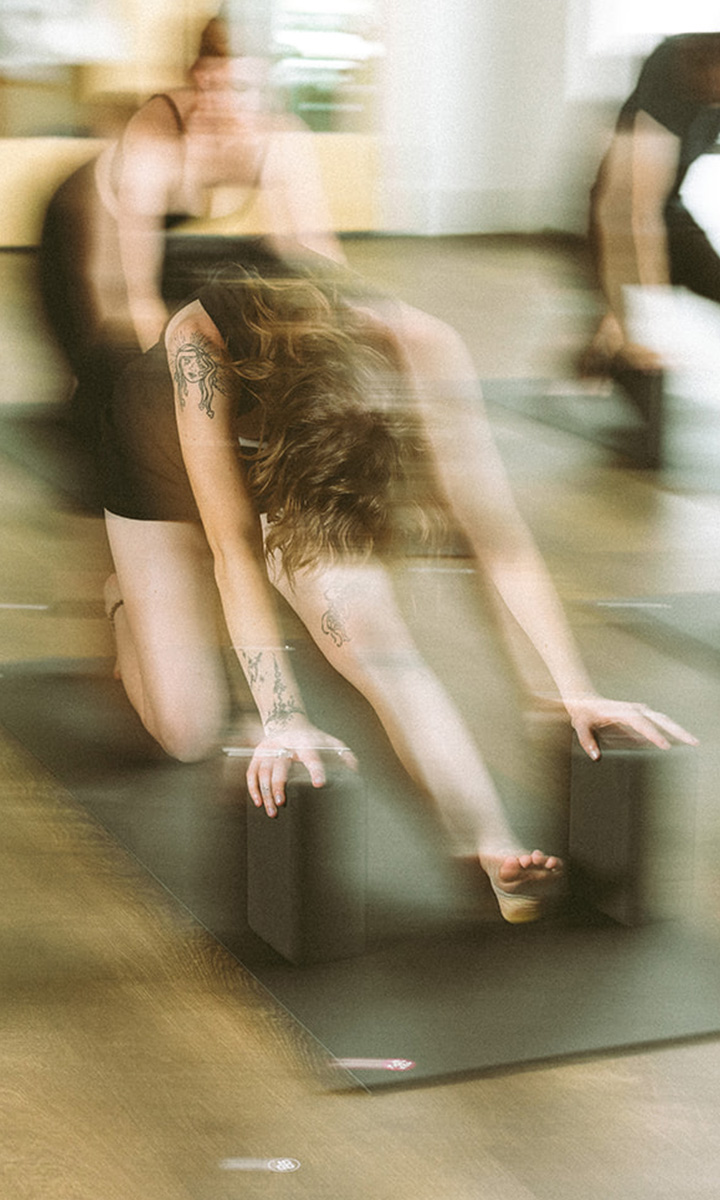 A person with tattoos practices a yoga stretch on a mat, using yoga blocks for support. The image has a blurred, dynamic effect, suggesting movement. Other people are visible in the background.