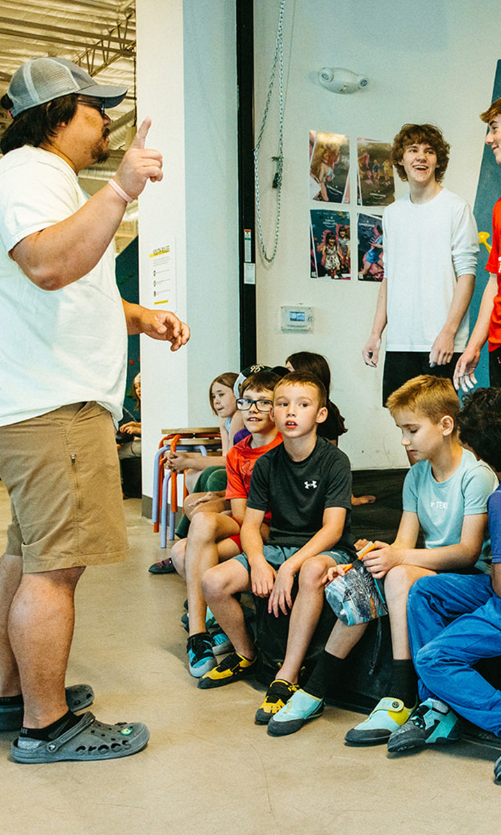 A man stands and gestures while addressing a group of seated children in a casual indoor setting. The kids listen attentively, and posters are visible on the wall behind them.