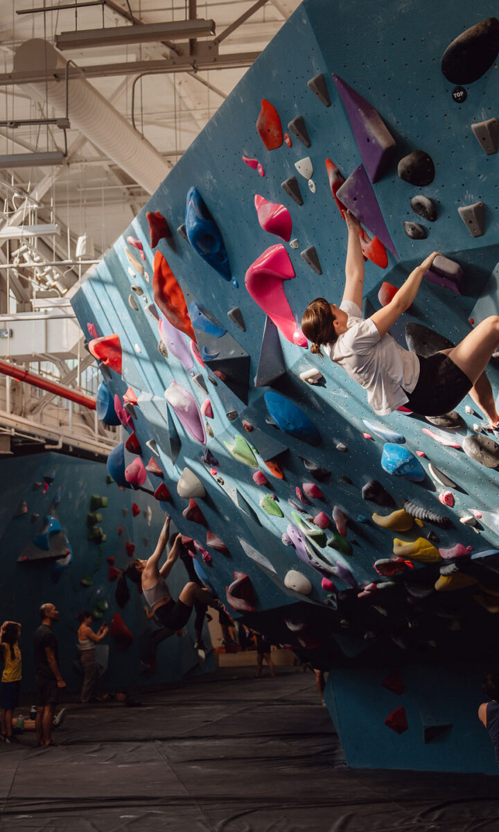 A group of people in a bright Brooklyn Climbing Yoga and Fitness gym, with one person scaling a blue climbing wall covered in colorful holds. Others watch or prepare to climb nearby as sunlight streams through large windows above.