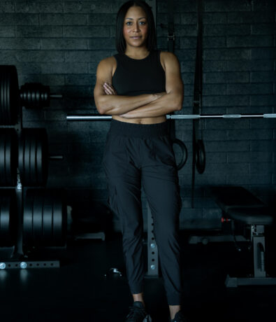 A woman in black athletic wear stands with arms crossed in a gym, in front of weightlifting equipment and a dark brick wall.
