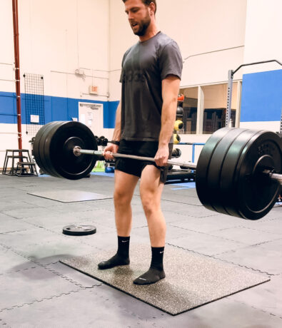 A man wearing a grey t-shirt, black shorts, and socks is performing a deadlift with a heavily loaded barbell in a gym setting. Gym mats cover the floor and equipment is visible in the background.