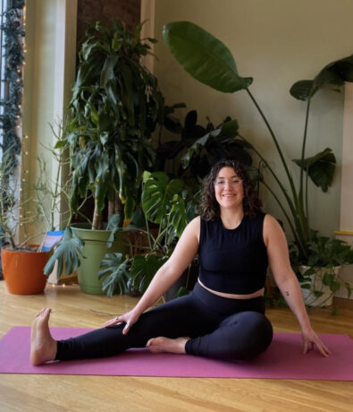 A woman in black workout clothes sits on a pink yoga mat indoors, stretching one leg out to the side and smiling. She is surrounded by large green houseplants and natural light from windows.