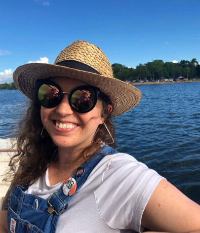 A woman wearing a straw hat, round sunglasses, and denim overalls smiles by a lake on a sunny day. Green trees and a blue sky with a few clouds are visible in the background.