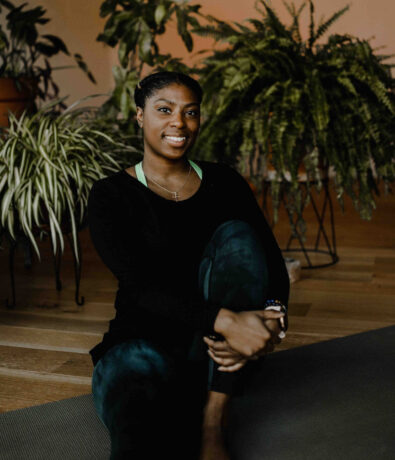 A woman sits on a yoga mat indoors, smiling at the camera, surrounded by various green houseplants. She wears a black long-sleeve top and dark leggings, with her arms wrapped around one bent knee.