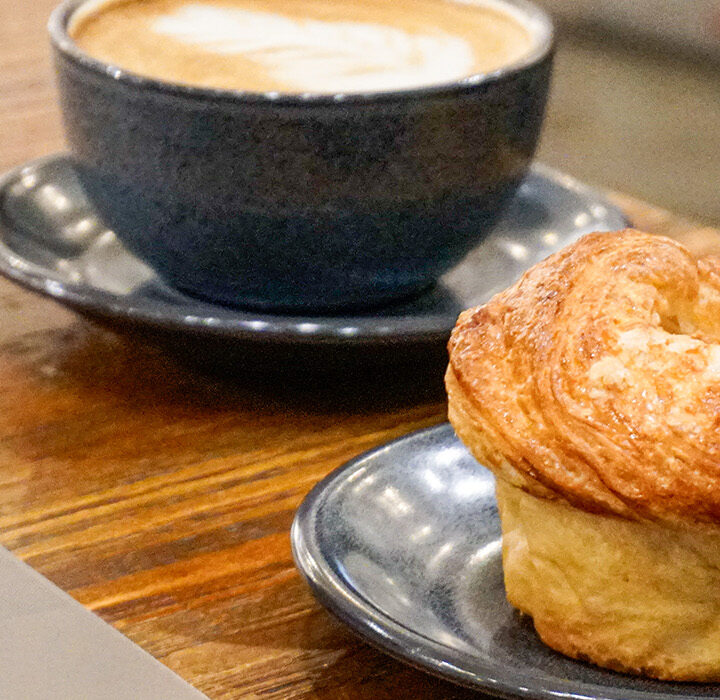 A blue coffee cup with latte art and a croissant muffin on separate blue plates sit on a wooden table at West Wall Cafe, next to a partially visible laptop.