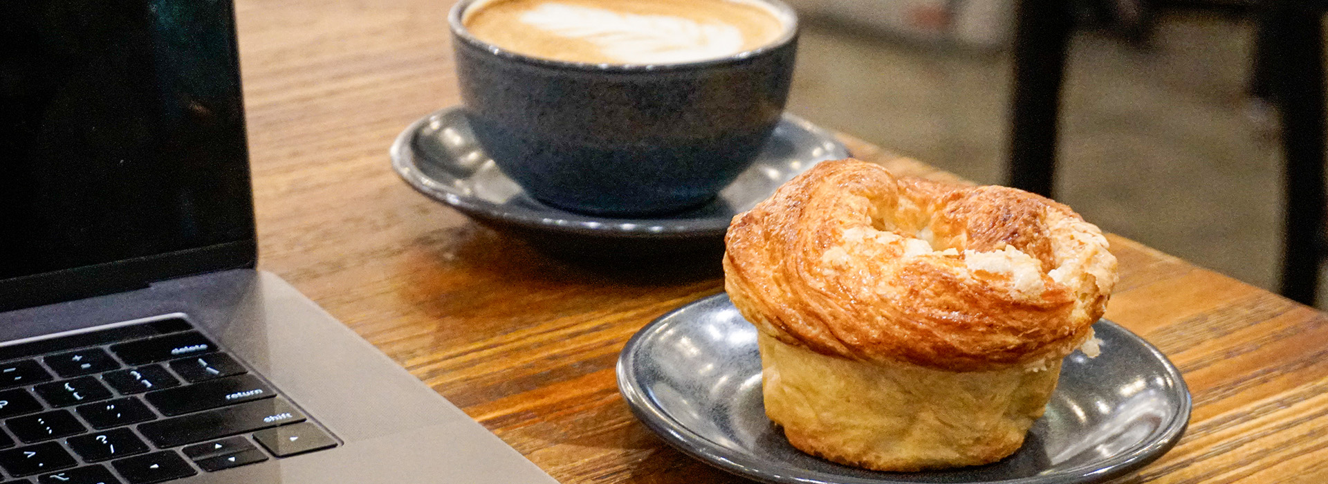 A blue coffee cup with latte art and a croissant muffin on separate blue plates sit on a wooden table at West Wall Cafe, next to a partially visible laptop.