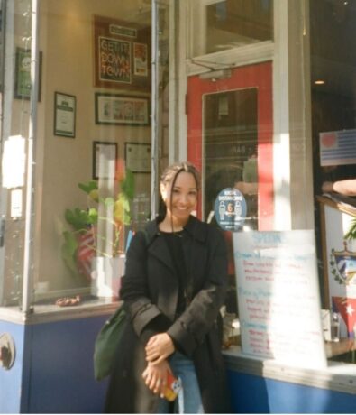 A woman with braided hair, wearing a black coat, stands smiling in front of a restaurant with a red, white, and blue door and climbing potted plants. A menu is posted outside, and framed posters are visible through the window.