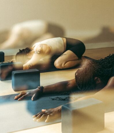 Two people stretch on yoga mats in a studio, reaching forward in a child’s pose with arms extended. Yoga blocks are placed beside them. The softly lit scene evokes the calm and focus found after an indoor rock climbing gym session.