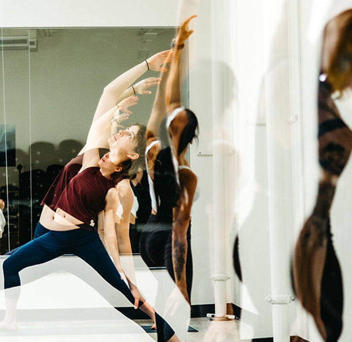 A group of people practicing yoga in a bright studio, stretching and reaching upwards before a mirror that reflects their movements—perfect for those enjoying the benefits of a summer membership.