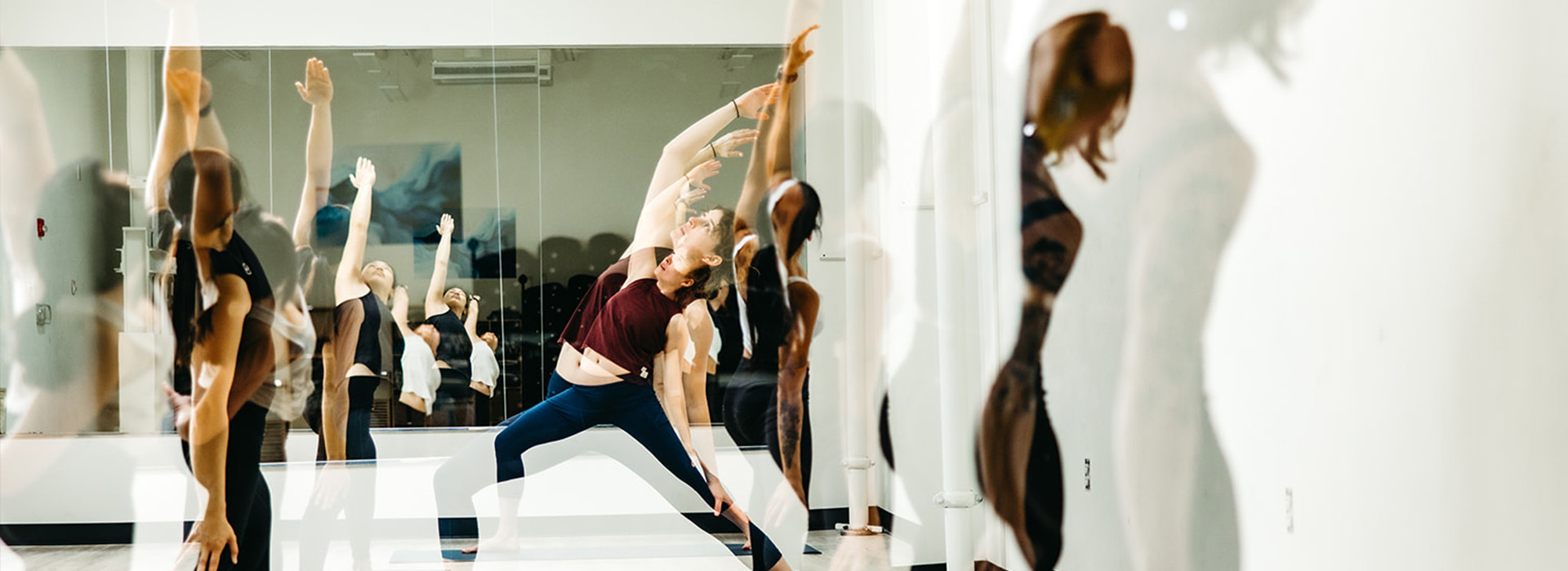 A group of people practicing yoga in a bright studio, stretching and reaching upwards before a mirror that reflects their movements—perfect for those enjoying the benefits of a summer membership.