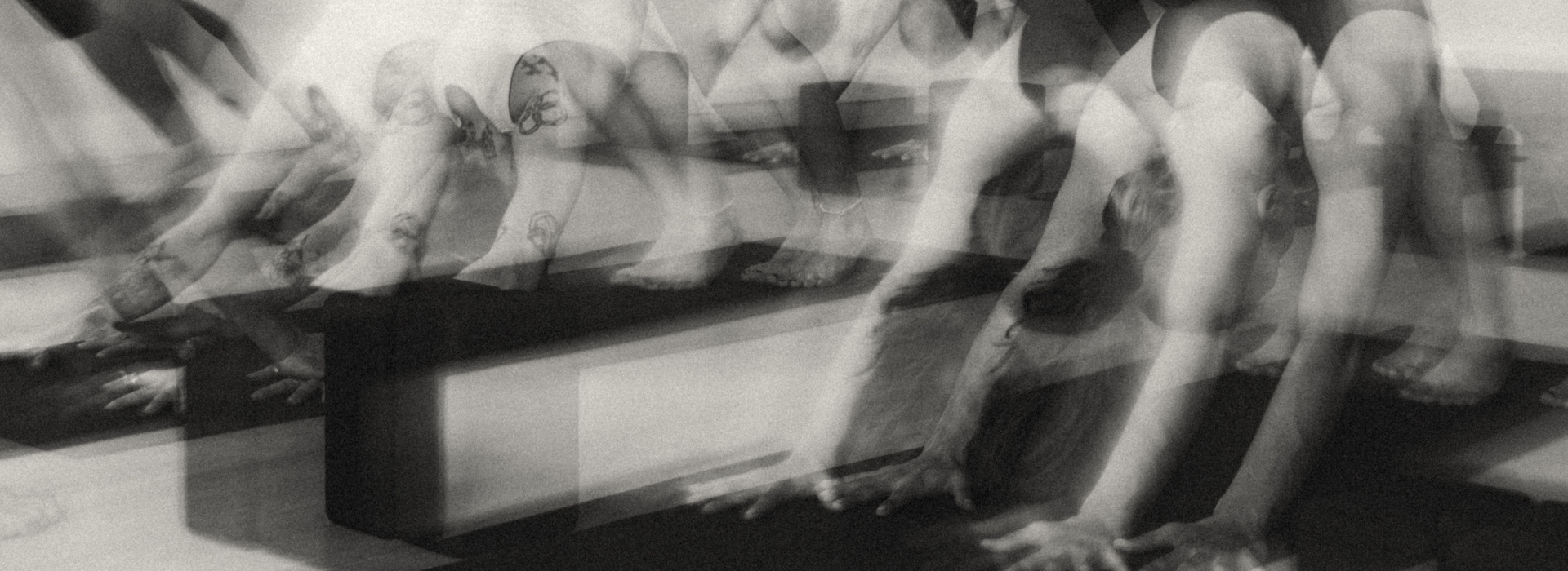 Black and white, blurred photo showing multiple overlapping images of tattooed arms and legs in a Vinyasa yoga pose on mats, creating a sense of movement and motion.