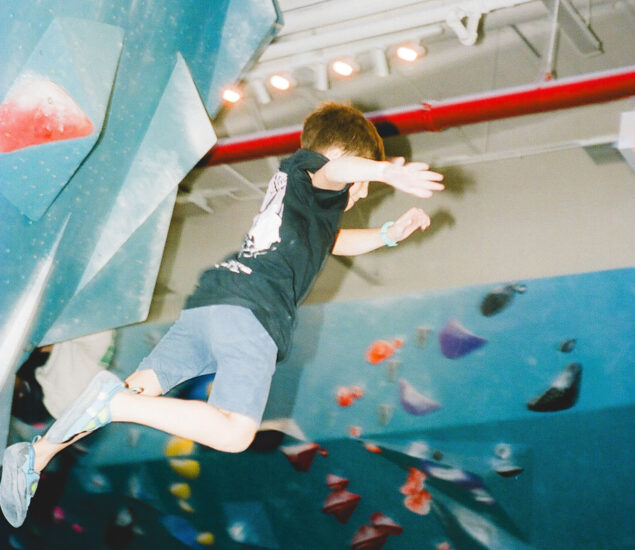 A child wearing a black shirt and blue shorts leaps mid-air off a climbing wall in an indoor Seattle Bouldering Climbing Gym, with colorful climbing holds visible in the background.