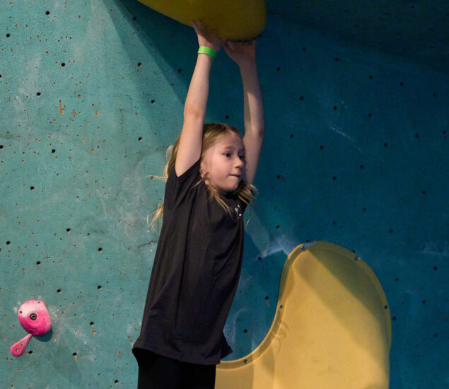 A young girl in a black shirt hangs by her arms from a yellow climbing hold on an indoor climbing wall at Seattle Bouldering Climbing Gym, looking focused and determined.