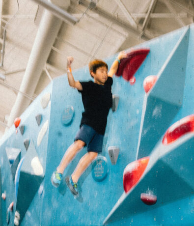 A young person in a black shirt and shorts jumps off an indoor climbing wall, arms raised, with colorful climbing holds visible on the blue wall behind them.