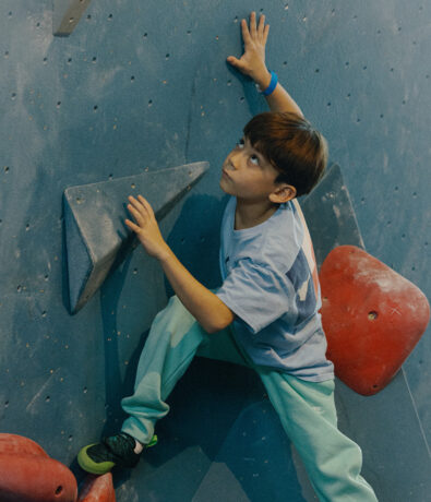 A young boy climbing an indoor rock wall looks up as he reaches for a handhold, wearing a light blue shirt and teal pants, with his foot on a red climbing hold.