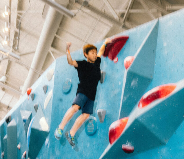 A young boy in a black shirt and blue shorts climbs an indoor rock climbing wall at a Seattle Bouldering Climbing Gym, gripping a red hold with one hand and lifting the other arm. The large, well-lit gym is perfect for After School Clubs.