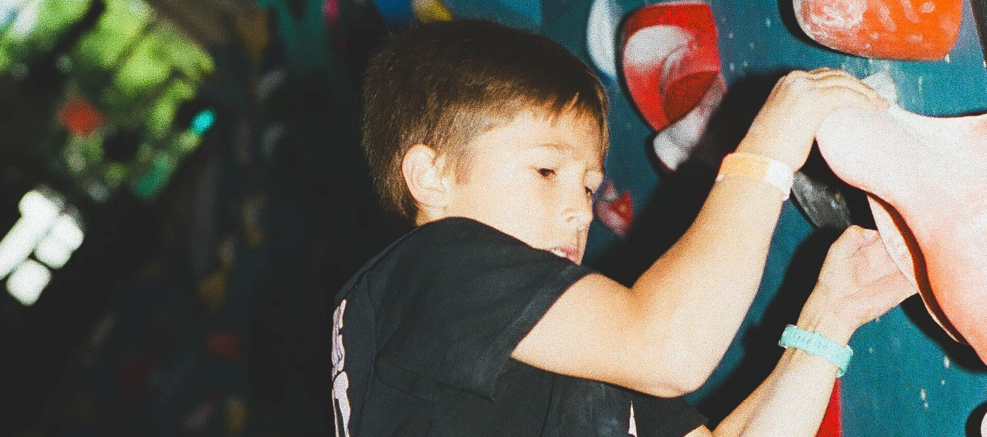 A young boy wearing a black shirt climbs an indoor rock climbing wall, gripping colorful handholds with both hands. Bright light highlights his focused expression during one of our exciting Youth Programs.