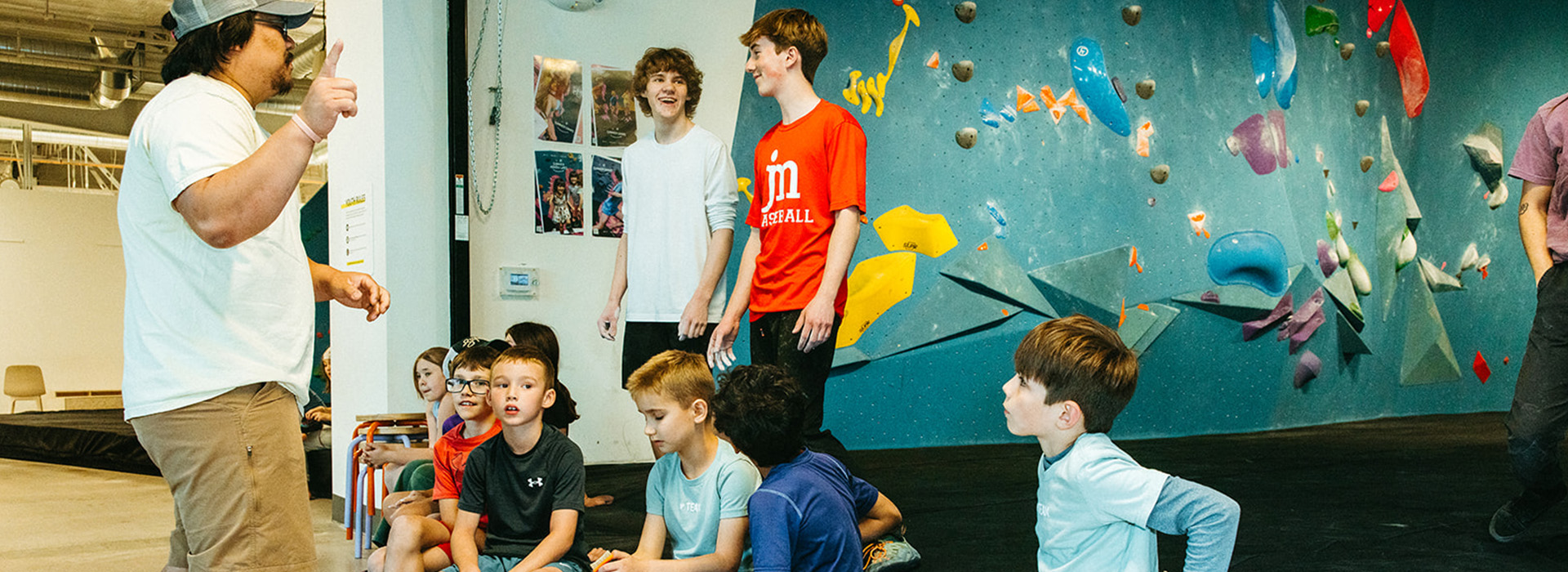 A group of children sit and stand near an indoor climbing wall while an adult gestures, possibly instructing them. The children appear attentive and engaged in a brightly lit gym setting.