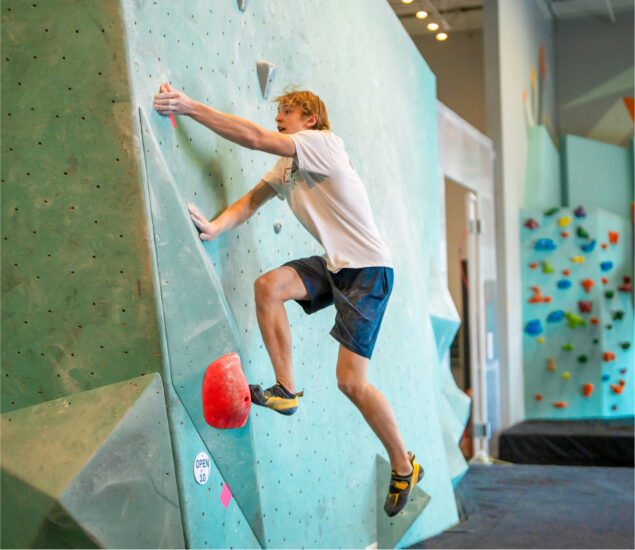 A man wearing a white t-shirt, shorts, and climbing shoes is bouldering on an indoor climbing wall, gripping holds with both hands and feet, focused on his ascent—perfect inspiration for youth development through challenging youth activities.