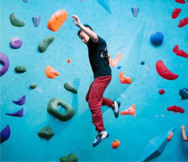 A person in red pants and a black shirt climbs a blue indoor bouldering wall with colorful handholds in orange, green, purple, and red. The person appears to be mid-jump or falling off the wall.