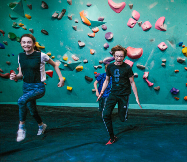 Two kids joyfully run toward the camera in a brightly colored indoor rock climbing gym, perfect for birthday parties, with a climbing wall and holds in the background.