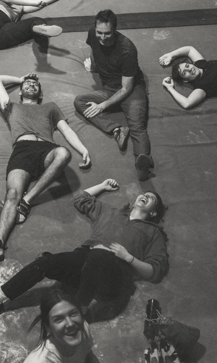 Five people are lying on the ground, laughing and smiling, suggesting they are relaxed and enjoying themselves. The image is in black and white, with a casual and joyful atmosphere.