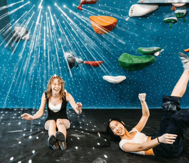 Two women laughing and sitting on the floor of an indoor climbing gym, colorful holds and bright, star-like light beams behind them. They're celebrating New Year 2025 with playful energy—don’t miss our promos available in all markets!.