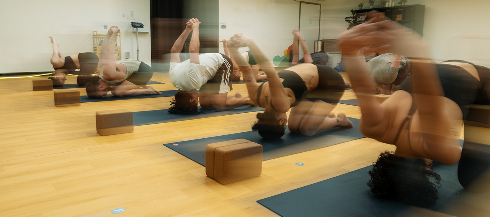 A group practices Hot Fusion Yoga on mats in a studio, performing a pose on their knees with arms clasped behind their backs. The image has a motion blur effect, and yoga blocks from Bouldering Project are placed beside each person.