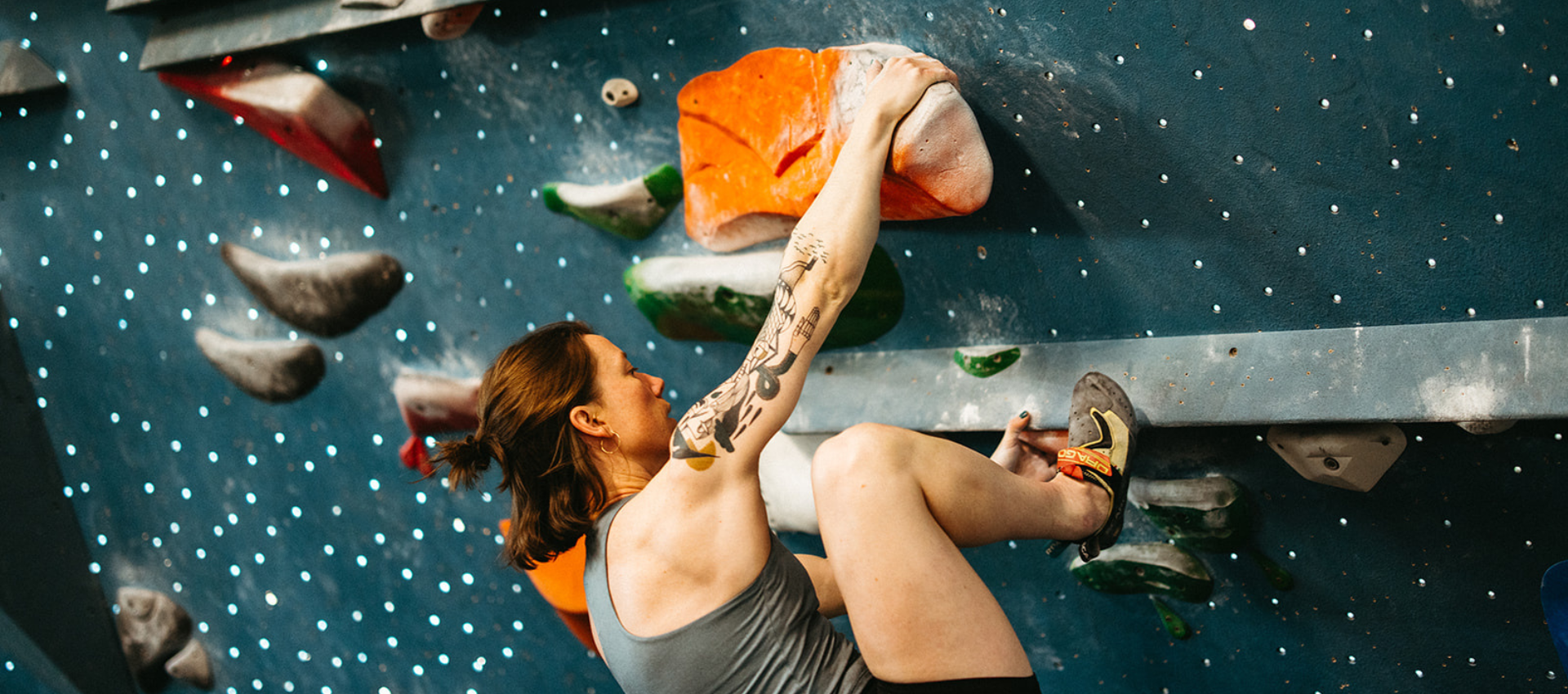 A woman with a tattoo on her left arm climbs an indoor bouldering wall, gripping a large orange rock climbing hold while wearing climbing shoes.