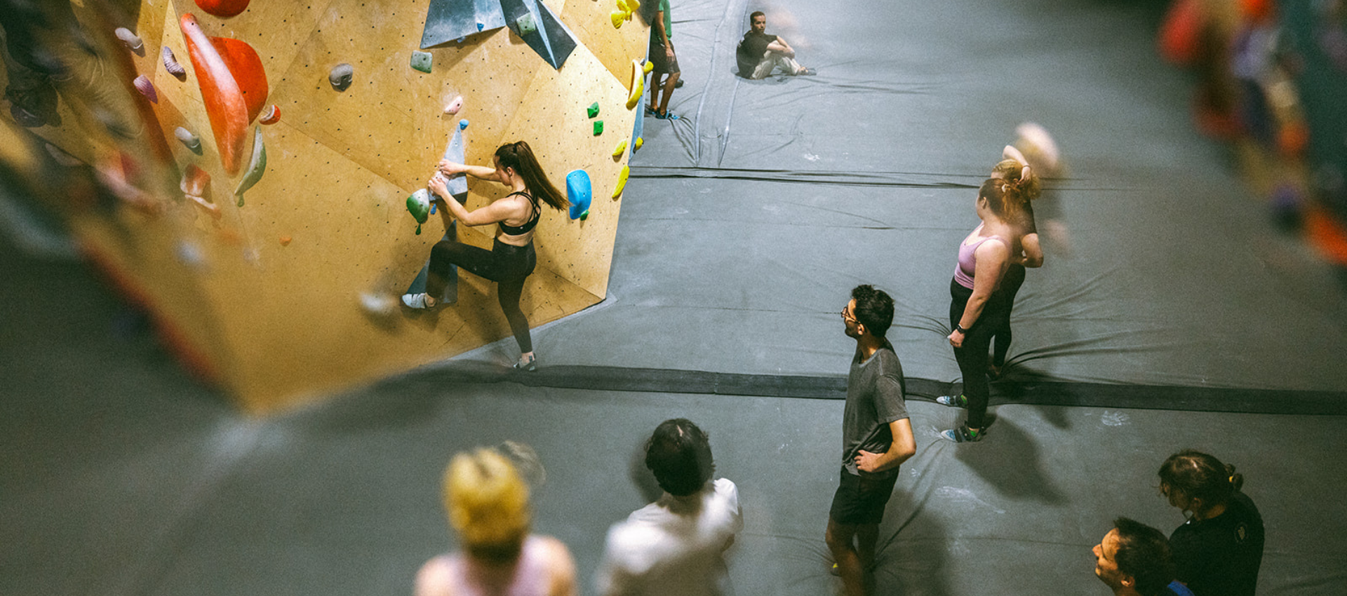 People inside a climbing gym watch as a woman scales a bouldering wall with colorful holds, tackling challenging climbing grades. Others stand or sit nearby, preparing to climb or observing. The atmosphere is active and social.