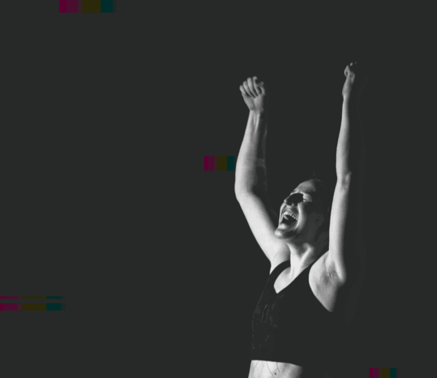 A person wearing a sports bra raises both arms triumphantly in the air, smiling and celebrating in dramatic lighting against a dark background at Boulderfest 2025.