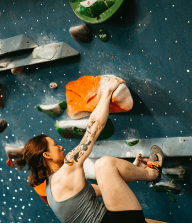 A person with a tattooed arm climbs an indoor bouldering wall, gripping vibrant rock climbing holds in orange and green, focused and wearing climbing shoes and a gray tank top.