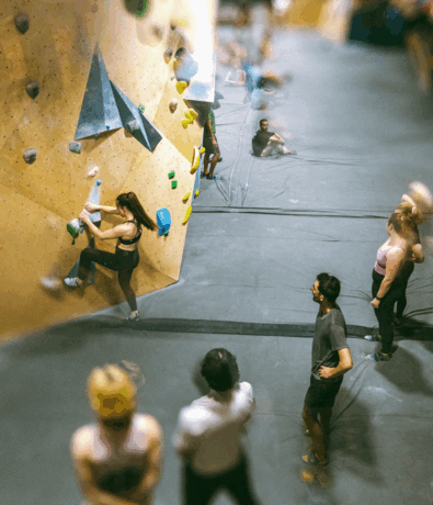 A group of people watch as a woman in climbing gear ascends an indoor bouldering wall with colorful holds, aiming for challenging climbing grades in a busy gym. Several others are seated or standing nearby, awaiting their turn.