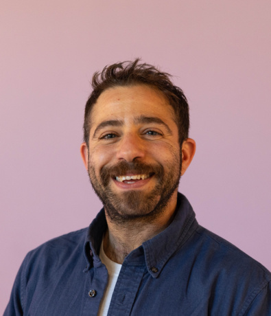 A man with short dark hair and a beard smiles at the camera, wearing a blue button-up shirt over a white t-shirt, standing in front of a plain light purple background, looking ready for an adventure at an Austin Climbing Gym.