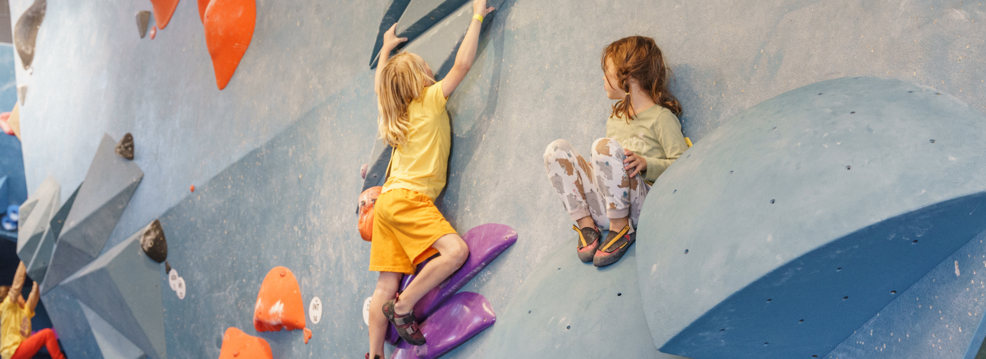 Two young children climb on a colorful indoor bouldering wall during one of the youth programs Boston offers. One, in yellow, climbs upward while the other, in patterned leggings, sits on a blue hold and looks toward their friend.