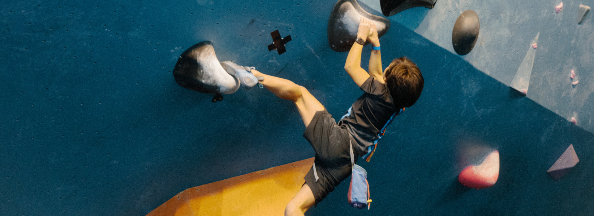 A young person wearing a harness climbs an indoor rock wall, gripping large holds with their hands and feet against a blue surface—an exciting scene from one of the top youth programs Boston has to offer.