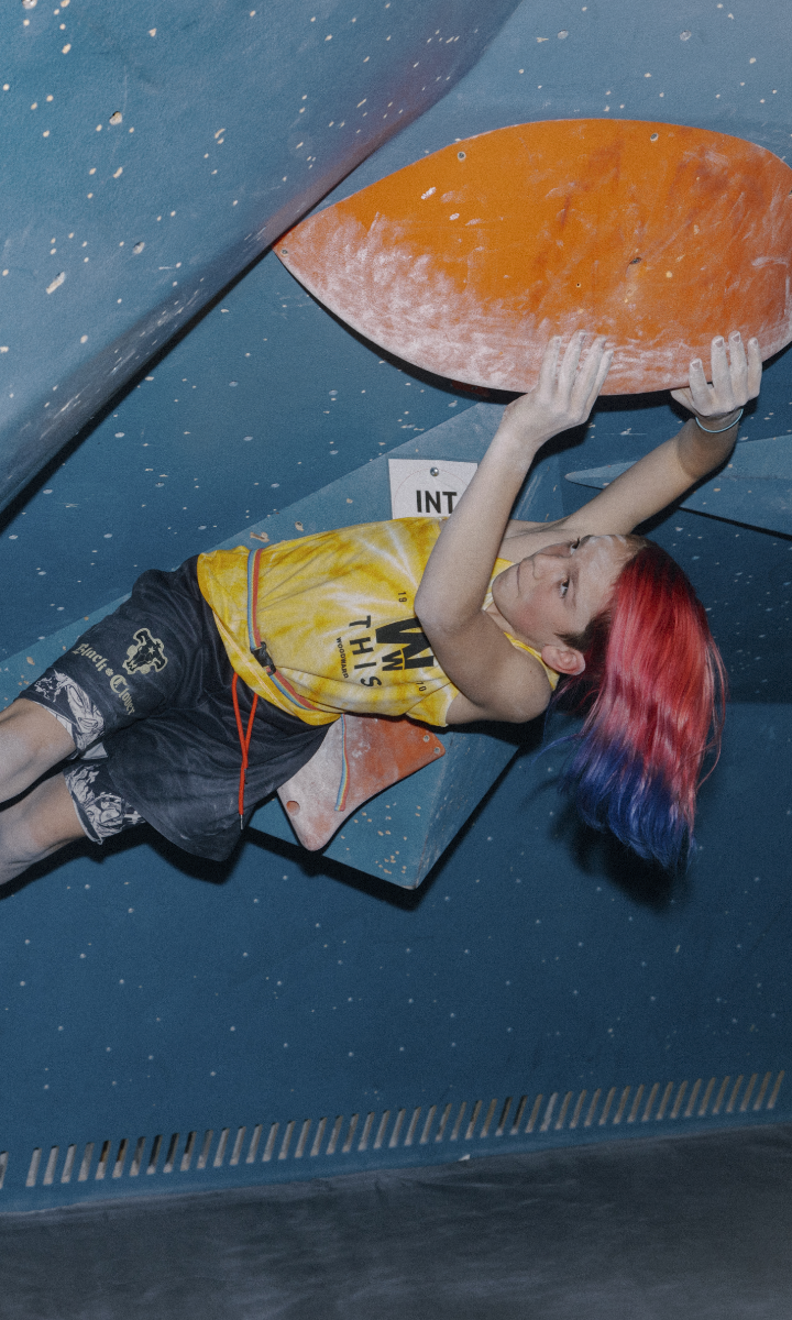 A young climber with red and blue hair hangs from an indoor bouldering wall at youth programs Boston, gripping a large orange hold. They wear a yellow tank top and black shorts, focused on the climb.