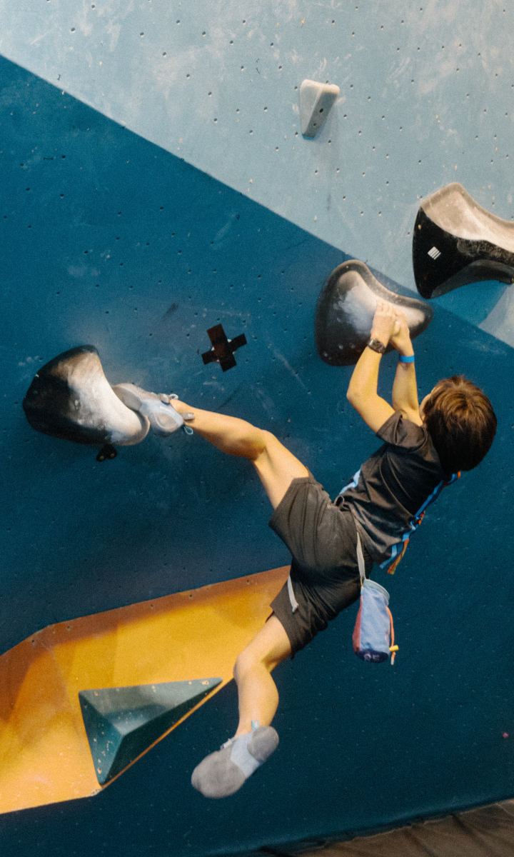 A young boy in a harness and climbing shoes grips large holds on an indoor climbing wall, reaching with one hand while balancing with his legs spread wide during one of the exciting youth programs Boston has to offer.