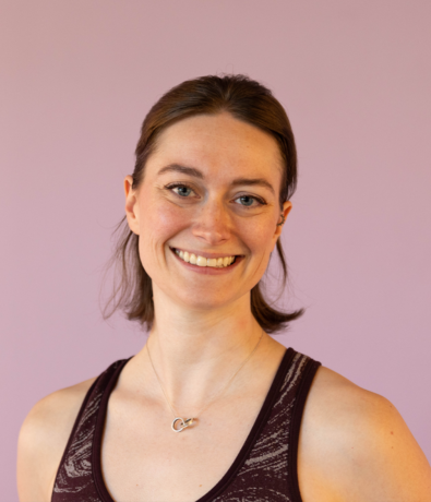 A woman with brown hair smiles at the camera, wearing a patterned burgundy tank top and a silver necklace. The solid light purple background gives a fresh vibe—perfect for a profile at your favorite Austin Climbing Gym.