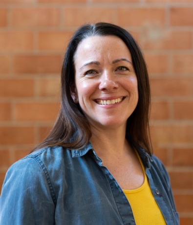 A woman with long brown hair smiles at the camera, wearing a yellow top and a blue denim shirt, standing in front of a red brick wall at an Austin Climbing Gym.